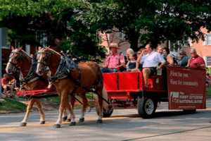 Two brown horses wearing harnesses pull a large parade float carrying many people. The side of the float has a banner with the First Citizens National Bank logo.