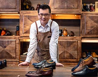 A man wearing an apron leans on a counter that is covered with new leather shoes