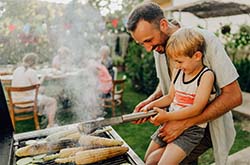 A man is holding a young boy happily. They are standing in front of a barbecue grill and the boy has utensils in his hand to help the man prepare the food that's on the grill.