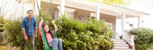 A father is pushing his young daughter on a swing in the yard in front of their home. They both look joyful and like they are having fun.