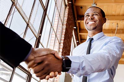 A man is smiling with his arm outstretched to shake another person's hand. Only the other person's hand is visible.